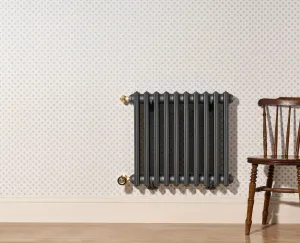 Grace 4 Column 660mm cast iron radiator in dark grey, wall-hung with Natural Brass valves, on dotted wallpaper beside a wooden chair.