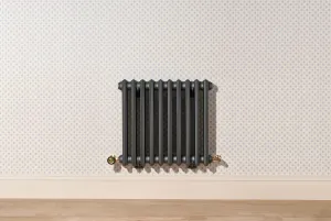 Wall-hung Grace 4 column cast iron radiator in dark grey Pencil with Natural Brass valves, on dotted wallpaper above a skirting and wooden floor.