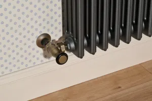 Close-up of a Grace 4 Column cast iron radiator in dark grey with a Natural Brass angled valve, against star-print wallpaper above a wooden floor.