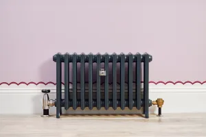Grace 4 Column cast iron radiator in Anthracite on feet, with a nickel angled valve and brass lockshield, against a lilac wall and pale wood floor.