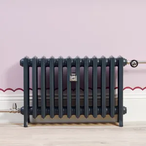 Grace 4 Column cast iron radiator in Anthracite on feet, with nickel valves and wall stay, against a lilac wall above a pale wood floor.