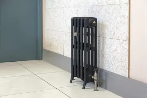 Grace 4 Column 660mm cast iron radiator in a dark finish on feet, with an antique brass valve, on a pale tiled floor against a tiled wall.