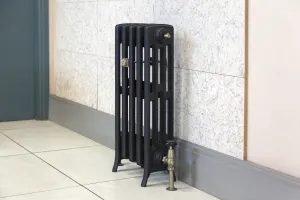 Short Grace 4 Column cast iron radiator in black on feet, with an antique brass angled valve, against a pale stone-effect wall on a tiled floor.
