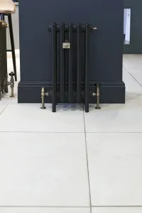 Narrow Grace 4 Column cast iron radiator on feet in a dark finish, with antique brass valves and wall stay, against a navy wall on pale tiles.