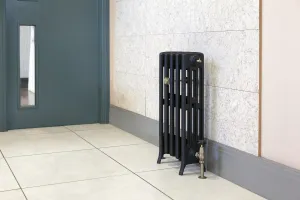 Grace 4 Column 660mm cast iron radiator in a dark finish on feet, with an antique brass valve, against a marble-effect wall on a tiled floor.