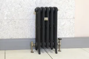 A five-section Grace 4 Column cast iron radiator in black on feet, with antique brass angled valves, against a marble-effect wall and grey skirting.