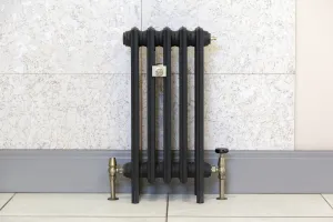 Grace 4 Column 660mm cast iron radiator in a dark matt finish, on feet with antique brass valves and wall stay, against a marble-effect wall.