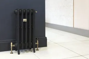 Grace 4 Column 660mm cast iron radiator in black on feet, with brass valves and wall stay, against a navy wall on a white tiled floor.