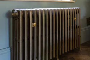 Grace 4 column cast iron radiator in Beaten Brass on feet with brass wall stays, under a window on a herringbone wood floor by a teal wall.