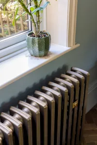 Grace 4 Column 660mm cast iron radiator in Beaten Brass beneath a white windowsill with a potted plant, against a pale blue wall.
