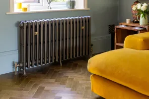 Grace 4 Column cast iron radiator in a metallic grey finish on feet under a window, with brass valves and stays, beside a yellow velvet sofa.