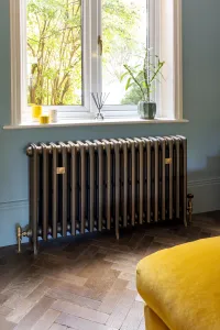 Plain Grace 4 Column cast iron radiator in a dark metallic finish on feet, brass valves and stays, under a window on herringbone parquet.