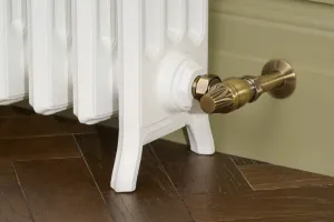 Close-up of a Grace cast iron radiator foot in Bright White with a Chatsworth angled thermostatic valve in antique brass, on herringbone oak.