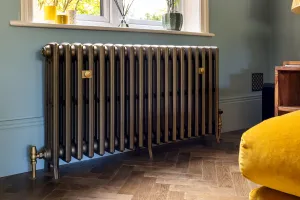 Grace 4 column cast iron radiator in Beaten Brass on feet beneath a window, with brass valves and wall stays on a herringbone oak floor.