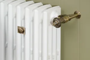 Close-up of a Grace 4 Column cast iron radiator in Bright White with an angled Chatsworth thermostatic valve in aged brass against a green panelled wall.