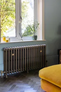 Grace 4 Column cast iron radiator in Beaten Brass on feet beneath a window, with brass valves, herringbone floor and a mustard sofa.