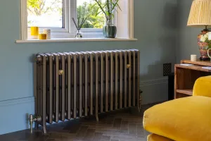 Grace 4 Column 660mm cast iron radiator in Beaten Brass on feet under a window, with brass stays and valves, by a yellow velvet sofa on herringbone floor.