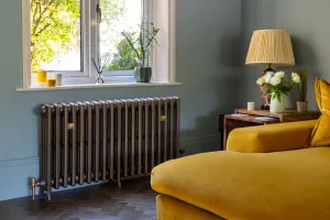 Grace 4 Column 660mm cast iron radiator in Beaten Brass on feet beneath a window, with brass wall stays and valve, beside a mustard velvet sofa.
