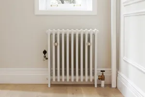 Grace 4 column cast iron radiator in Ceramic White on feet beneath a window, with brass wall stays and valves, on a light oak floor.