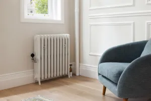 Grace 4 Column cast iron radiator in Ceramic White on feet below a window, with wall stays, valves, oak floor and a blue armchair.
