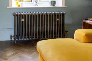 Grace 4 column cast iron radiator in Beaten Brass on feet under a window, with angled valves, herringbone floor and a yellow velvet sofa.