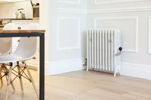 Grace 4 column cast iron radiator in Ceramic White on feet with nickel valves, against panelled walls beside a kitchen dining table.
