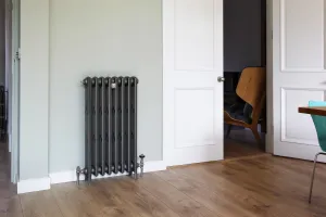 Grace 4 Column 960mm cast iron radiator in Soft Pewter on feet, with nickel valves, against a pale green wall beside a white door on a wooden floor.
