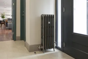 Tall Grace 4 Column cast iron radiator in a metallic pewter-style finish on feet, with chrome valves, beside a dark grey glazed door on a tiled floor.