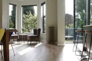 A tall Grace 4 Column cast iron radiator in Soft Pewter on a pale wall beside a bay window, with an armchair and oak floorboards.