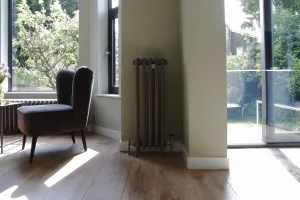 A tall, narrow Grace 4 Column cast iron radiator in Soft Pewter on feet, with angled valves, beside glass doors and an oak floor.