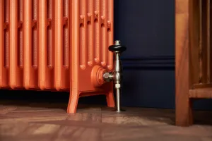 Close-up of a Grace 6 Column cast iron radiator in orange on feet, with a satin nickel angled valve, navy wall and parquet floor.