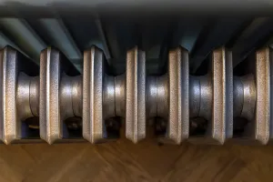 View down onto the tops of a Grace cast iron radiator's sections in a Beaten Brass metallic finish, above a herringbone wood floor.