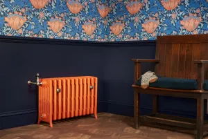 Orange Grace 6 column cast iron radiator on feet with a nickel valve, against a navy dado wall below floral wallpaper, beside a wooden settle.
