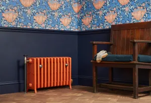 Orange Grace 6 column cast iron radiator on feet with a chrome valve, against navy panelling below floral wallpaper, beside a wooden settle.