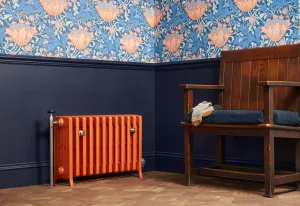 Grace 6 column cast iron radiator in orange on feet with a nickel valve, beside navy panelling, floral wallpaper and a wooden settle.