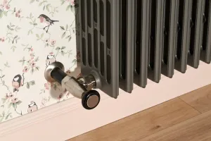 Close-up of a Grace 6 Column cast iron radiator in Quartz Grey with a silver-toned Chatsworth angled valve, by bird-print wallpaper and a wooden floor.