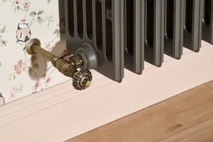 Close-up of a Grace 6 Column 485mm cast iron radiator in Quartz Grey with a Natural Brass Chatsworth valve, by floral wallpaper and pink skirting.