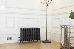 Grace 6 Column cast iron radiator in Astral Grey Dark on feet, with angled valves, against panelled white walls on herringbone oak flooring.