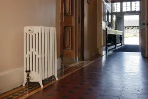 A plain white Grace 6 column cast iron radiator on feet with a nickel valve, in a hallway with a red and black tiled floor.
