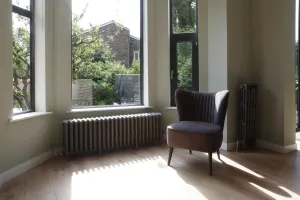 Grace 6 column cast iron radiator in Soft Pewter on feet beneath a bay window, with a tall slim radiator beside a grey armchair on an oak floor.