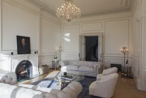 A low dark grey cast iron radiator under a panelled wall beside double doors, in a drawing room with tufted sofas, a fireplace and chandelier.