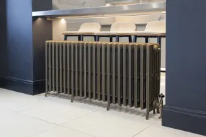 Grace 6 Column 660mm cast iron radiator on feet in a bronze-toned finish with angled valve, beneath a kitchen counter with bar stools.