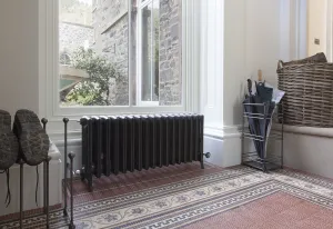 Grace 6 Column cast iron radiator in Astral Grey Dark on feet under a window, on a mosaic tiled floor beside an umbrella stand and boot rack.