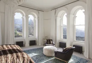 Four low Grace cast iron radiators in Astral Grey Dark beneath tall arched windows in a white bedroom with armchairs, a fur pouffe and a blue rug.