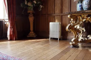 White Grace 6 Column 660mm cast iron radiator on feet with valves, against oak panelling beside a gilded console table on a wooden floor.