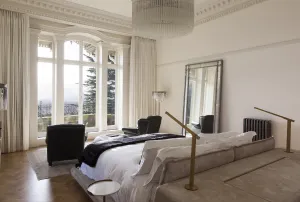 A Grace cast iron column radiator in a dark metallic finish against a white wall behind a low bed, with tall arched windows and herringbone floor.