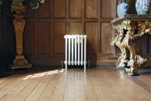 A white Grace 6 Column cast iron radiator on feet with brass valves, against oak panelling on a wooden floor beside a gilded console table.