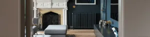 Grace 4 column cast iron radiator in dark blue-grey on a dark blue wall, beside a white carved fireplace, herringbone floor and grey ottoman.