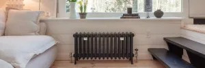 Grace 4 Column 500mm cast iron radiator in Farrow & Ball Black Blue No. 95 on feet, with chrome valves, under a window beside a white sofa.