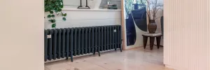 Long, low Grace four-column cast iron radiator in Farrow & Ball Black Blue No. 95 on feet, below a white panelled wall on a pale timber floor.